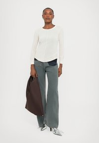 Woman wearing white long-sleeve shirt, wide-leg jeans, silver pointed heels, holding large dark brown tote bag, standing against white background.