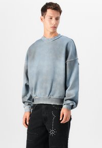 Sweatshirt de gola redonda azul claro com uma superfície desbotada e texturizada e com bainha canelada. Usado com jeans pretos desgastados com detalhes de tachas.