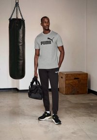 Grå Puma t-skjorte med svart logo, mørke joggebukser og svarte joggesko. Holder en svart treningsbag; punching bag i bakgrunnen.