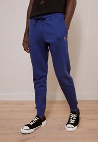 Blue cotton sweatpants with elastic waistband and drawstring. Features side pockets and a small embroidered logo. Paired with black sneakers.