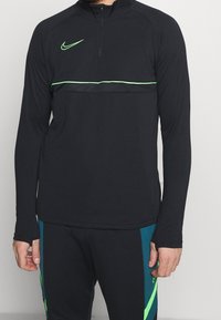 Musta pitkähiha urheilupaita vetoketjukauluksella, vihreillä yksityiskohdilla ja Nike-logolla rinnassa. Paidassa on vaakasuora vihreä raitakuosi.