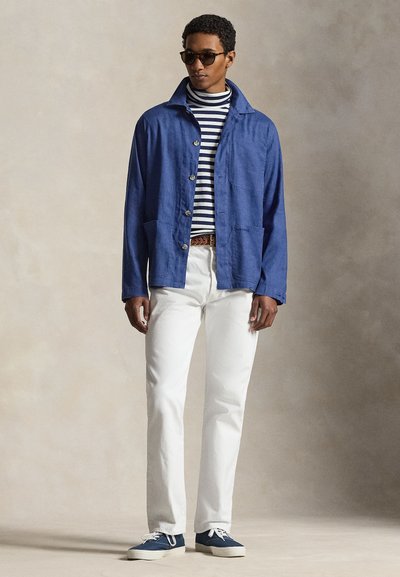 Άνδρας που φοράει σκούρα γυαλιά ηλίου, μπλε μπουφάν, ριγέ ζιβάγκο, άσπρο παντελόνι και μπλε αθλητικά παπούτσια σε απλό μπεζ φόντο.