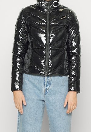 Shiny black quilted puffer jacket with front zipper, high collar with white text, worn with light blue denim jeans.