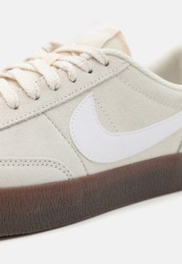 Beige Wildlederschuh mit einem weißen Lederlogo, strukturierten braunen Gummisohle und cremefarbenen Schnürsenkeln. Hat ein kleines Logo auf der Zunge.