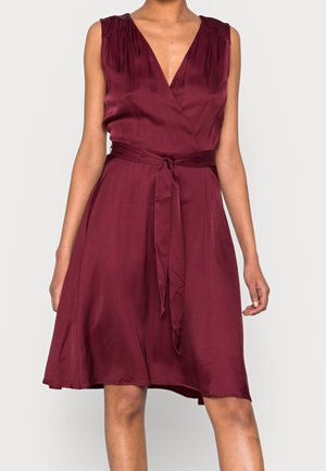 Bez rękawów bordowa satynowa sukienka z dekoltem w serek i paskiem w talii, do kolan, lekko rozkloszowana, noszona przez stojącą osobę.