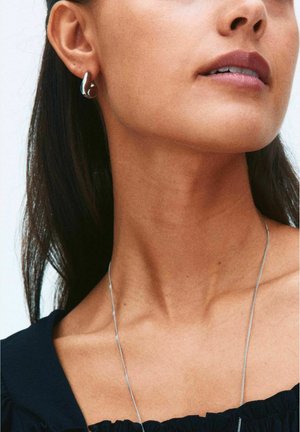 Lähikuva naisen alahampaista, kaulasta ja olkapäästä, joka käyttää hopeisia renkkikorvakoruja ja ohutta hopeista kaulakorua mustan topin kanssa.