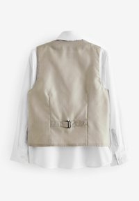 Gilet beige à motif avec une texture lisse, doté d'un détail de boucle en argent à la taille, porté par-dessus une chemise blanche à col.