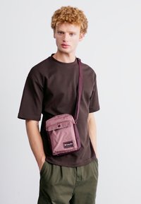 Borsa a tracolla in nylon viola con chiusura a patta, tracolla regolabile e patch con logo, indossata sopra una maglietta scura e pantaloni cargo.