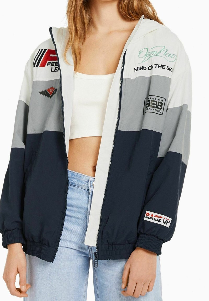 Ung person iført en vindjakke i tre farver med racer-relaterede mærker, hvid crop top og lyseblå jeans, stående med hænderne afslappede.