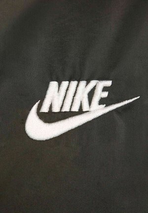 Logo "NIKE" ricamato in bianco su uno sfondo di tessuto nero testurizzato, con un accento a forma di swoosh che si estende dalla lettera 'K'.