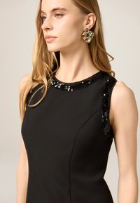 Sort kjole med en figursyet silhuet, der har en halsudskæring og skulderdekorationer pyntet med palietter. Modellen har bølget hår og gyldne øreringe.