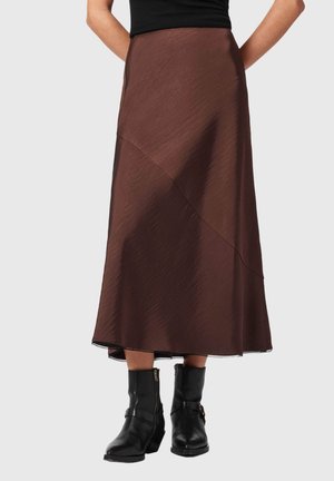 Personne portant une jupe midi marron avec des coutures diagonales et des bottines noires, se tenant devant un fond clair uni.
