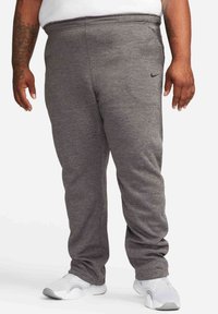 Homme portant un pantalon de survêtement Nike gris et des chaussures de sport blanches, debout devant un fond blanc.