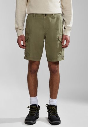 Olive green cargo shorts with a button closure, side pockets, and a small logo patch. Worn with a light sweater and black boots.