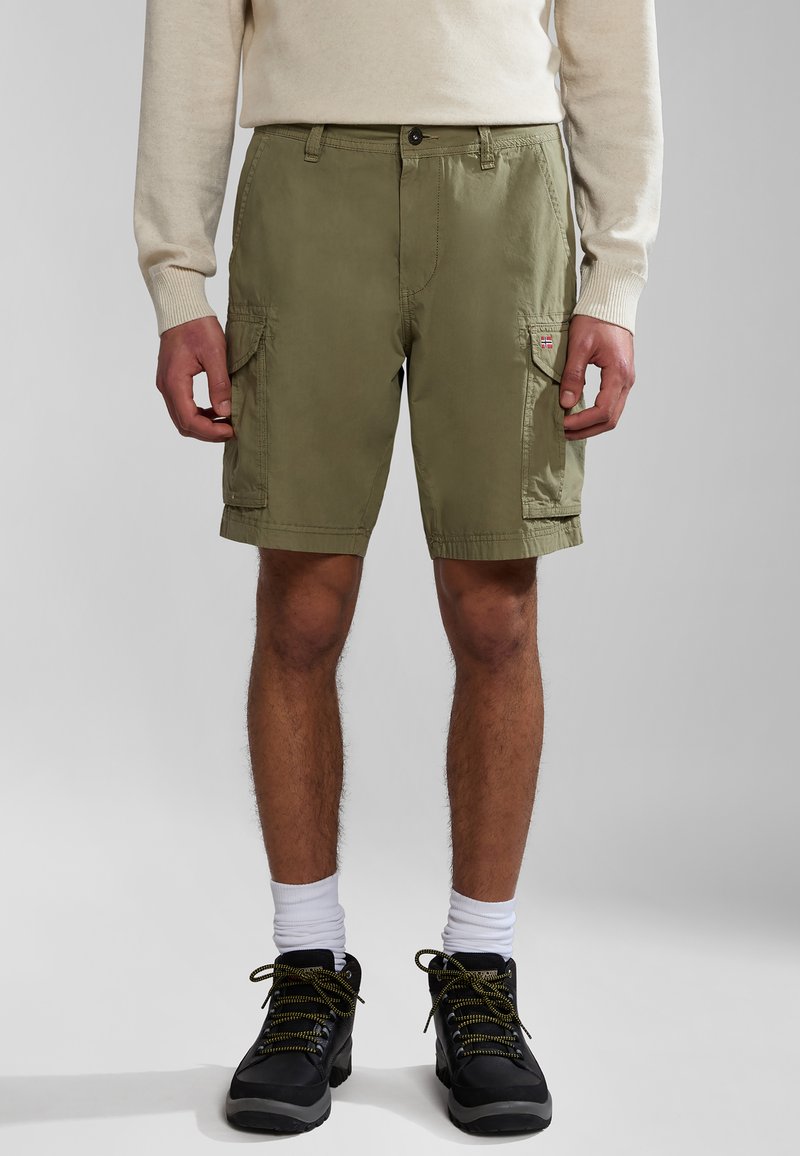 Pantalones cortos cargo color verde oliva con cierre de botón, bolsillos laterales y un pequeño parche con el logo. Llevado con un suéter ligero y botas negras.