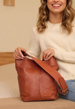 Bruine leren tote bag met een zachte textuur, brede opening en verstelbare schouderriem, voorzien van een logo-patch aan de voorkant.