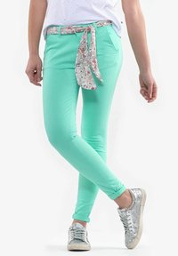 Mintgröna figursydda byxor med ett blommigt mönstrat knytbälte. Har en slät textur och uppvikt fåll. Matchas med sneakers i silverglitter.