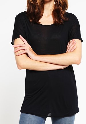 Frau mit schulterlangem braunem Haar, trägt ein schwarzes T-Shirt mit kurzen Ärmeln und blaue Jeans, die Arme vor der Brust verschränkt.