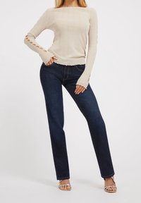 Top beige a coste in maglia con maniche lunghe e ritagli, abbinato a jeans a gamba dritta in denim scuro. L'insieme è completato da sandali argentati con cinturini.