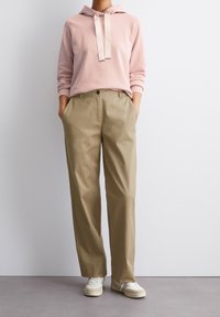 Felpa rosa chiaro con tasca anteriore e coulisse, abbinata a pantaloni beige a gamba dritta. Sneakers bianche completano l'outfit.