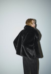 Sort jakke i imiteret pels med høj krave, firkantet form og blød tekstur. Båret over mørke jeans mod en lysegrå baggrund.