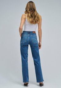 Mujer con cabello largo y ondulado, vistiendo una camiseta sin mangas blanca, vaqueros de cintura alta azul y sandalias de tacón negro, de pie contra un fondo gris plano.