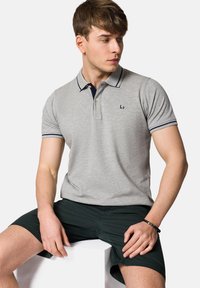 Szary polo z granatowym kołnierzykiem i akcentami na rękawach; zawiera trzy guziki i haftowane logo na piersi. Spodenki są ciemnozielone.