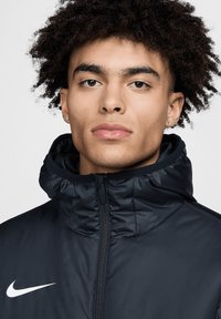 Navyblaue gepolsterte Jacke mit hohem Kragen, durchgehendem Reißverschluss und weißem Nike-Logo auf der linken Brust; glatte Textur und leichtes Design.
