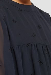 Tmavě modré šaty s texturovanou tkaninou, které mají průsvitné rukávy a vyšívaný vzor. Plisovaná tkanina v oblasti pasu dodává objem.