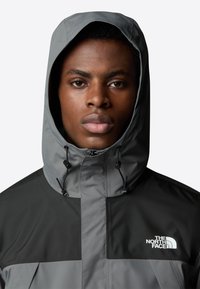 Grijze en zwarte jas met een capuchon, voorzien van een rits aan de voorkant, verstelbaar trekkoord en het "The North Face" logo op de linkerkant van de borst.