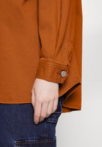 Chemise surdimensionnée orange rouille avec poignet boutonné, tissu texturé et détails de couture visibles, portée avec un pantalon en denim bleu foncé.