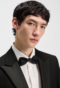 Noor mees, kellel on lühikesed tumedad juuksed, seljas must smokingujakk, valge särk ja must kikilips, taustal hele ühevärviline taust.