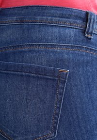 Närbild på en bakficka av blå denimjeans med orange sömmar och en röd skjorta instoppad i midjan.