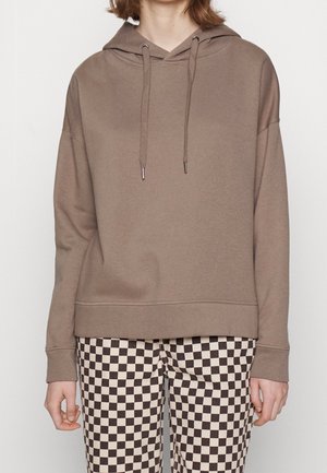 Personne portant un sweat à capuche taupe avec des cordons et un pantalon à carreaux noirs et blancs, debout devant un fond uni.