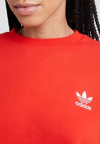 Červená mikina s kulatým výstřihem a žebrovaným límcem. Na levé části hrudi se nachází bílé logo Adidas se třemi listy. Měkká textilie.