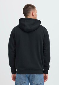 Sort hættejakke med en stor hætte, lavet af blødt materiale. Har ribbede manchetter og kant, med en afslappet pasform. Bagudsnittet viser minimal detaljer.