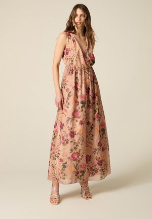 Frau trägt ein ärmelloses, V-Ausschnitt, knöchellanges Blumenkleid mit pinken, roten und orangen Blumen, kombiniert mit Riemchen-Sandalen mit hohen Absätzen.