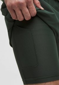 Grüne Sportshorts aus strukturiertem Material, mit einer Seitentasche, genähten Nähten und einem enganliegenden Design. Der Stoff ist dehnbar und leicht.