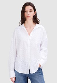 Weiße Bluse mit Knopfleiste, Lochmuster, langen Ärmeln und kragenlosem Ausschnitt. Getragen über hellblauen Denim-Jeans.