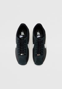 Nike sportcipők fekete bőr felsőrésszel, zöld velúr részletekkel, fehér swoosh logóval, lapos fekete fűzőkkel és kerek orrú kialakítással.