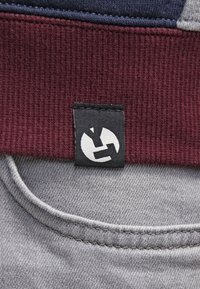 Svart klädesetikett med vitt abstrakt logotyp på vinröd ribbad tyg ovanför grått sömtäckt material.