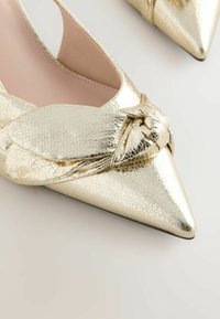 Chaussures plates dorées à bout pointu avec une finition texturée et un détail nœud décoratif sur le devant.