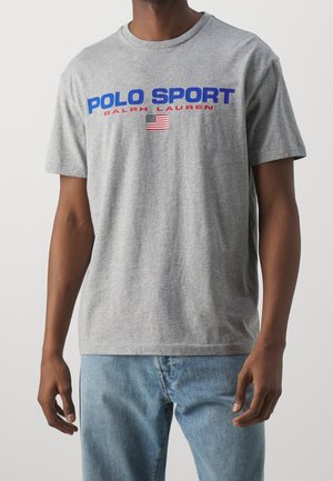 Mand iført en lysegrå t-shirt med teksten "Polo Sport Ralph Lauren" og et lille amerikansk flag, kombineret med lyseblå jeans.