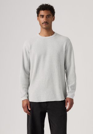 Homme aux cheveux bouclés et moustache portant une chemise en maille gaufrée gris clair à manches longues et un pantalon noir, debout devant un fond uni.