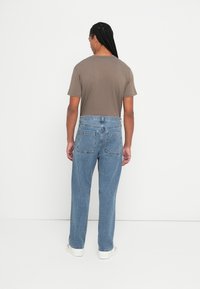 Άνδρας με πλεχτά μαλλιά, φοράει ένα καφέ t-shirt, ανοιχτό γαλάζιο τζιν και άσπρα αθλητικά παπούτσια, στέκεται μπροστά σε έναν άσπρο τοίχο.