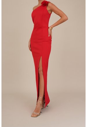 Femme portant une robe rouge à une épaule avec un détail floral et une fente haute à la cuisse, associée à des sandales dorées à talons hauts à brides.