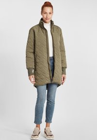 Quiltēta olīvu zaļa jaka ar augstu apkakli, rāvējslēdzēja priekšu un ribotiem roku galiem. Valkāta pāri baltam turtleneckam un ziliem džinsiem ar tenisēm.