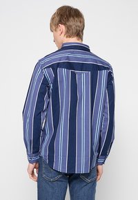 Camicia da uomo a maniche lunghe con righe verticali blu e azzurro. Presenta un colletto button-down e un orlo dritto con pieghe laterali.