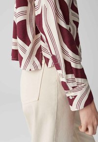 Camicia a maniche lunghe a fantasia bordeaux e crema con polsini abbottonati, abbinata a pantaloni di jeans chiari con tasche posteriori visibili.