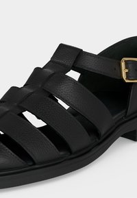 Μαύρο δερμάτινο σανδάλι ψαρά με χρυσή αγκράφα και πολλαπλές λωρίδες πάνω από το πόδι και κατά μήκος των πλευρών.